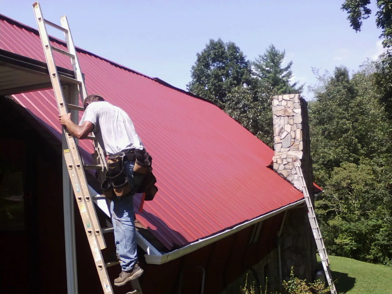Roofer on a ladder beside a red metal roof and stone chimney — Soffit & Fascia Repair by Roofing Brothers in Loris